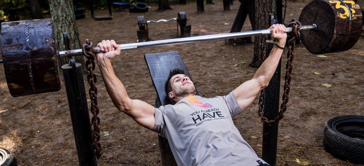 Man weightlifting outdoors, using a bench press setup amid trees for strength training.